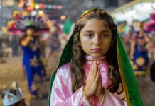 A young girl with hands clasped in prayer wears a pink and green costume evoking the traditional outfit of the Virgin of Guadalupe during a Fiestas Guadalupanas procession, a highlight of traditional Puerto Vallarta December 2025 events in the downtown district.