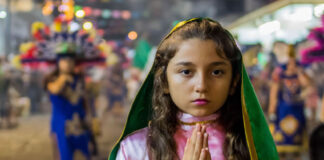 A young girl with hands clasped in prayer wears a pink and green costume evoking the traditional outfit of the Virgin of Guadalupe during a Fiestas Guadalupanas procession, a highlight of traditional Puerto Vallarta December 2025 events in the downtown district.