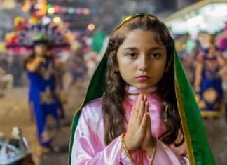 A young girl with hands clasped in prayer wears a pink and green costume evoking the traditional outfit of the Virgin of Guadalupe during a Fiestas Guadalupanas procession, a highlight of traditional Puerto Vallarta December 2025 events in the downtown district.