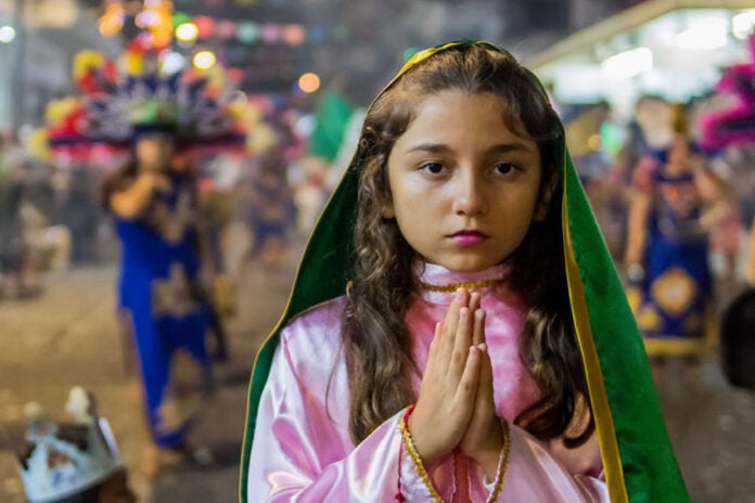 A young girl with hands clasped in prayer wears a pink and green costume evoking the traditional outfit of the Virgin of Guadalupe during a Fiestas Guadalupanas procession, a highlight of traditional Puerto Vallarta December 2025 events in the downtown district.