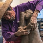 Artisan and sculptor Jose Garcia Antonio of Oaxaca, Mexico, in his studio leans over a large piece of clay he is sculpting into the shape of a human figure. He's wearing a purple button down shirt and a traditional cloth sombrero.