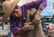 Artisan and sculptor Jose Garcia Antonio of Oaxaca, Mexico, in his studio leans over a large piece of clay he is sculpting into the shape of a human figure. He's wearing a purple button down shirt and a traditional cloth sombrero.