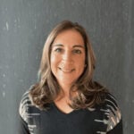 Headshot of a smiling woman, artist Tamanna Bembenek. She is standing in a black sweater with a black-and-white geometric pattern on the shoulders. She has long light-brown hair. She is standing against a dark gray textured background.