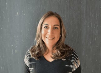 Headshot of a smiling woman, artist Tamanna Bembenek. She is standing in a black sweater with a black-and-white geometric pattern on the shoulders. She has long light-brown hair. She is standing against a dark gray textured background.