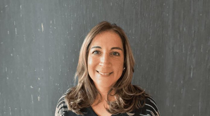 Headshot of a smiling woman, artist Tamanna Bembenek. She is standing in a black sweater with a black-and-white geometric pattern on the shoulders. She has long light-brown hair. She is standing against a dark gray textured background.