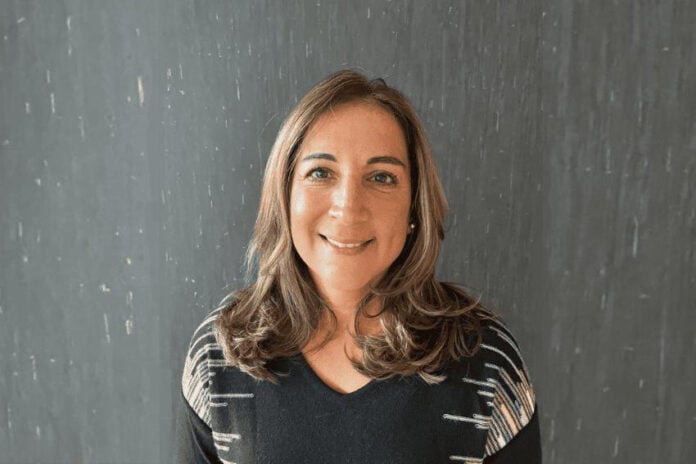 Headshot of a smiling woman, artist Tamanna Bembenek. She is standing in a black sweater with a black-and-white geometric pattern on the shoulders. She has long light-brown hair. She is standing against a dark gray textured background.