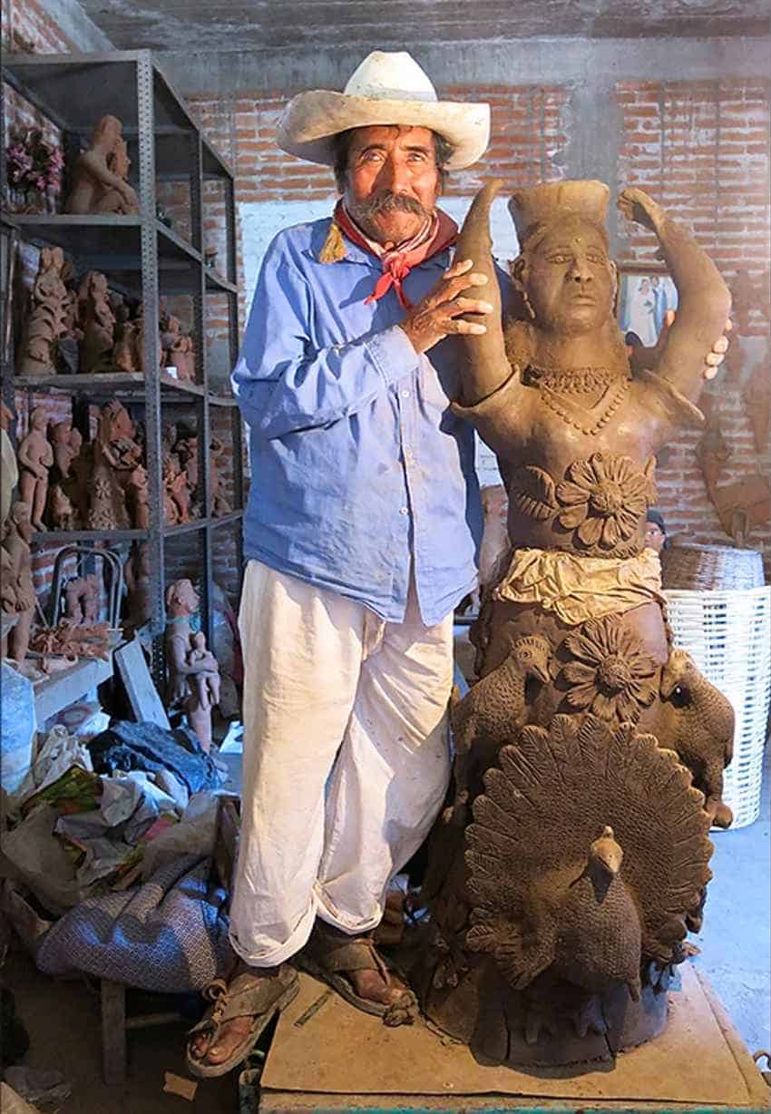 Mexican asulptor Jose Garcia Antonio stands in his redbrick-lined Oaxaca workshop. He's posing with a brown life-sized clay sculpture of an older stocky woman in traditional dress with her arms raised. The sculpture is behind another shorter clay sculpture of three peacocks.