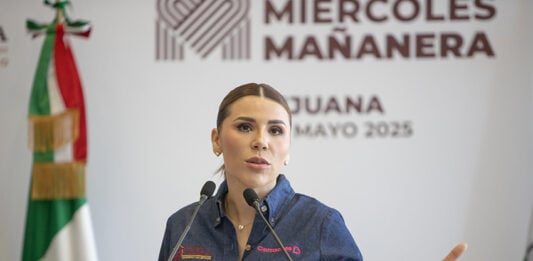 Woman speaking at a press conference with Mexican flag in background.