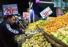 A customer selects fruit in a market with prices displayed in Mexican pesos