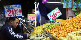 A customer selects fruit in a market with prices displayed in Mexican pesos