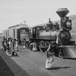 A vintage black-and-white photograph from the Porfiriato era, featuring Mexican campesinos in white and with straw hats and an elderly woman carrying a large ceramic cistern in front of a Ferrocarril Central Mexicano steam locomotive at a rural station in Mexico during the nation's Porfiriato era, which followed Porfirio Díaz's rise to power under the Plan of Tuxtepec.