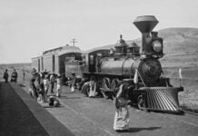 A vintage black-and-white photograph from the Porfiriato era, featuring Mexican campesinos in white and with straw hats and an elderly woman carrying a large ceramic cistern in front of a Ferrocarril Central Mexicano steam locomotive at a rural station in Mexico during the nation's Porfiriato era, which followed Porfirio Díaz's rise to power under the Plan of Tuxtepec.