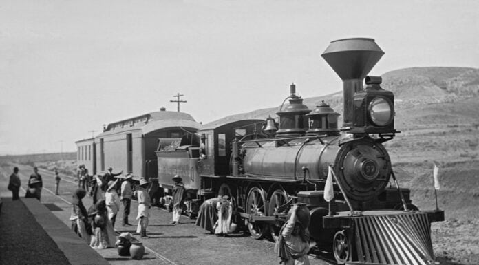 A vintage black-and-white photograph from the Porfiriato era, featuring Mexican campesinos in white and with straw hats and an elderly woman carrying a large ceramic cistern in front of a Ferrocarril Central Mexicano steam locomotive at a rural station in Mexico during the nation's Porfiriato era, which followed Porfirio Díaz's rise to power under the Plan of Tuxtepec.