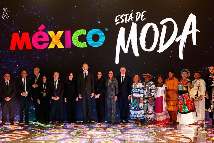 Politicians in black suits and Indigenous Mexicans in colorful tradition clothing stand on a stage under the words "México está de moda"