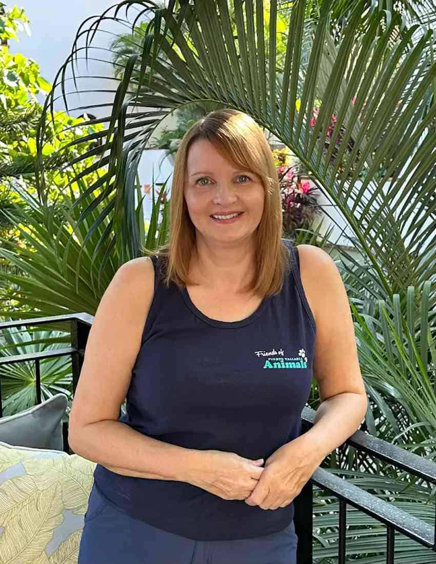 Melanie Henderson of Puerto Vallarta smiling on a balcony, wearing a Friends of Puerto Vallarta Animals tank top standing in front of a deck chair and a the giant leaf of a palm tree.