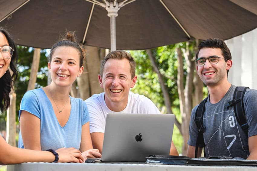 A group of American students at Tec de Monterrey in Monterrey, Mexico. They are sitting at an outdoor table that has a Mac laptop and a backpack. They are smiling.