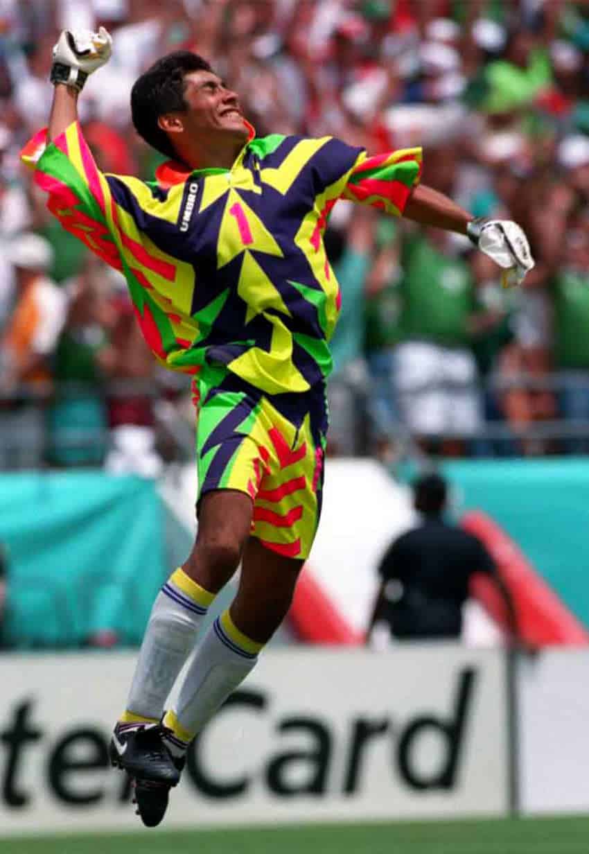 Mexico soccer player Jorge Campos at a game in the 1994 World Cup. He is caught in the middle of a joyful jump with one fist in the air, wearing a kit of neon green, neon pink, yellow, and blue. and white blue and yellow socks