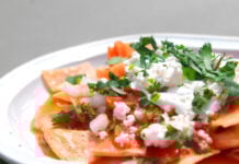 A close-up shot of a plate of red chilaquiles, a staple authentic Mexican recipe, topped with fresh crema, crumbled queso fresco, diced onions and cilantro.