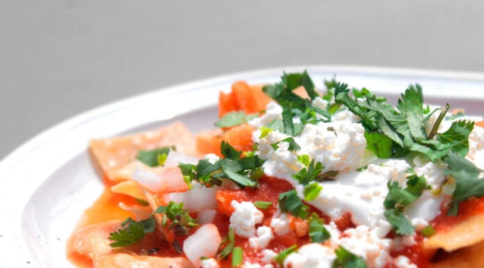 A close-up shot of a plate of red chilaquiles, a staple authentic Mexican recipe, topped with fresh crema, crumbled queso fresco, diced onions and cilantro.