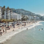 Aerial image of Puerto Vallarta beach with people in the water and countless beach umbrellas all over the beach. There are white adobe apartment buildings lining the coast in the background.