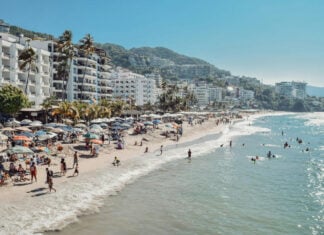 Aerial image of Puerto Vallarta beach with people in the water and countless beach umbrellas all over the beach. There are white adobe apartment buildings lining the coast in the background.