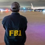 A man in an FBI jacket watches a plane on a runway