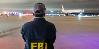 A man in an FBI jacket watches a plane on a runway