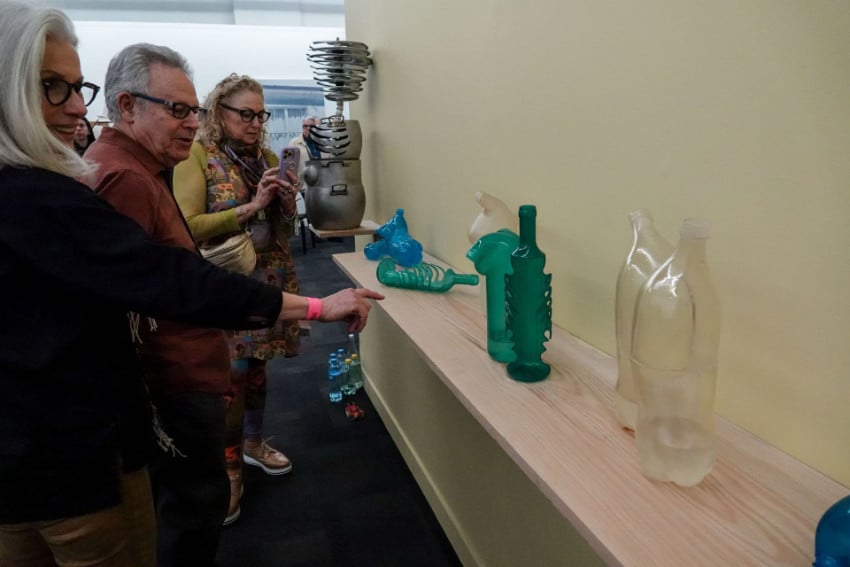 A group of three art enthusiasts, all middle-aged, in a gallery setting examines contemporary glass sculptures during Mexico City Art Week 2025, including translucent green and bottle installations displayed on a light wooden shelf against an undecorated beige wall. A middle-aged woman in the foreground points toward the recycled-material art pieces while nearby, another women in her group takes a photo with a smartphone.