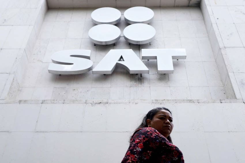 A Mexico City woman in a floral shirt and long dark hair walking past the offices of Mexico's SAT tax collection agency, a white cantera building. She is staring at the photographer with a grim expression.