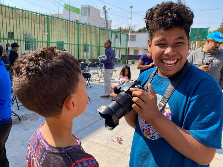 Two Latin American migrant boys smiling and laughing as one holds a Sony digital SLR camera to take photos of his friend. They are in a migrant holding center in Mexico City.
