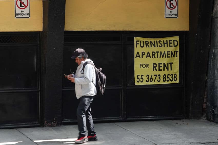 A man wearing a baseball cap and backpack is walking on a sidewalk while looking at his smartphone, passing by a yellow vinyl sign in English pinned to the outside of a building. The sign advertisesfurnished apartments for rent and a Mexico City phone number to call.