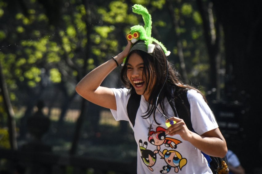 A Mexican teenaged girl wearing a Powerpuff Girls tee shirt in white and wearing a headband feature a green monkey with yellow sunglasses. She is laughing at something off camera and is in a park in Mexico City