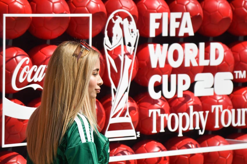 A young blonde woman in a green Mexico national team jersey with red sunglasses on her her head stands in front of a red Coca-Cola and FIFA World Cup 26 Trophy Tour backdrop made of soccer balls.