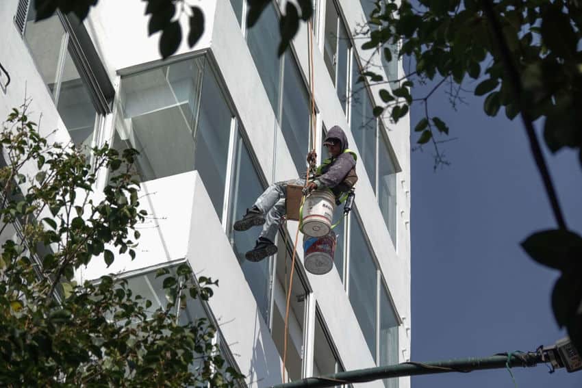 A Mexican man sits on two industrial sized paint buckets with a wooden board on top, a makeshift scaffold attached to ropes. The man is lowering himself down a multistory building and looking down at the ground below.