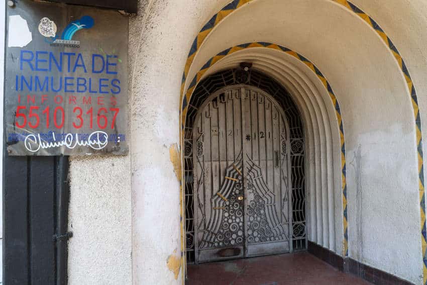 A weathered arched doorway with a decorative wrought iron gate in Mexico City, featuring a 'Renta de Inmuebles' sign and a phone number, illustrating the local rental market,