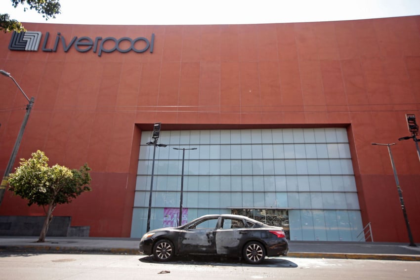 A burned-out black car with white scoring in various parts of it from being set on fire. It sits in front of the glass doors of a massive facade of a Liverpool department store entrance in Guadalajara, Mexico