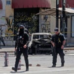 Police patrolling on a Guadalajara street with a burned-out sedan in the background.