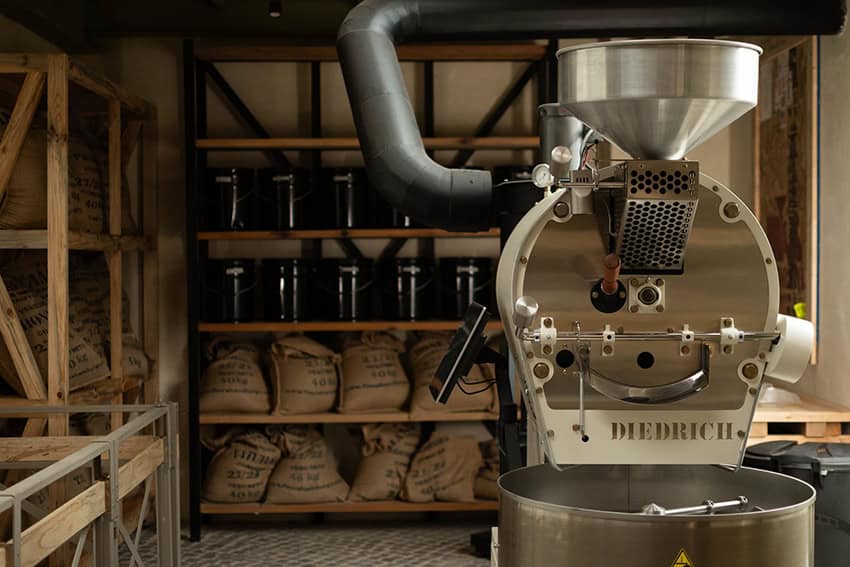 A large coffee roasting machine with bags of coffee and bottles stored in the background