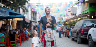 A man holds a baby and a child's hand standing on the street of a Mexican town