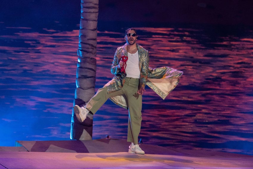 Bad Bunny putting on a show at the Estadio Azteca in Mexico City in 2022. He is wearing green cotton pants and a white tank top undershirt with a sequined jacket in light greens and pinks. Behind him is a trunk of a fake palm tree and a projected image of water.