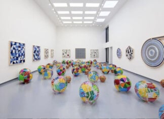 A spacious white gallery room filled with a contemporary installation for Mexico City Art Week, featuring dozens of colorful, patterned inflatable spheres scattered across a polished gray floor. The surrounding white walls are adorned with various geometric and abstract artworks, including blue-toned modular paintings and a large circular spiral piece, illuminated by a grid of bright skylights.