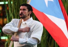Musician Bad Bunny in a white jacket and wearing an onstage headset/microphone clutches a life-sized Puerto Rican flag as part of his Super Bowl 2026 performance. Behind him are fake green stalks of sugar cane.