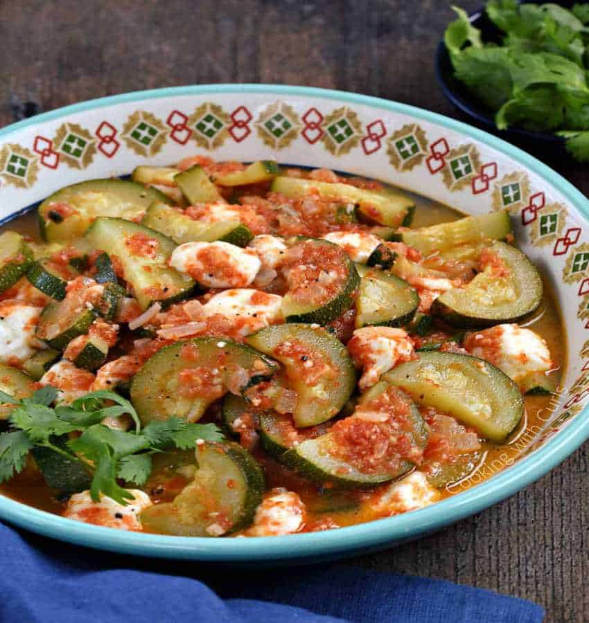 A decorated white bowl with light-blue trim is filled with Mexican squash with cheese, a sautéed zucchini and tomato dish with melted panela cheese.