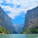 A wide-angle view of the towering limestone walls of Sumidero Canyon in Chiapas, Mexico, rising above the Grijalva River under a blue, cloudy sky.