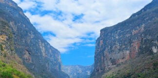 A wide-angle view of the towering limestone walls of Sumidero Canyon in Chiapas, Mexico, rising above the Grijalva River under a blue, cloudy sky.