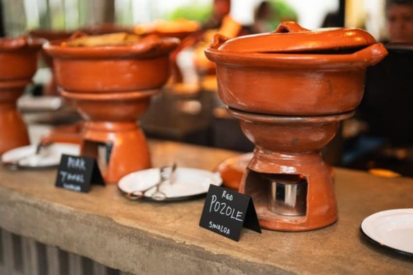 Close-up of large clay cooking pots with lids, labeled "Red Pozole Sinaloa" and "Pork Tamal," sitting on individual warming stands on a rustic wooden buffet table.