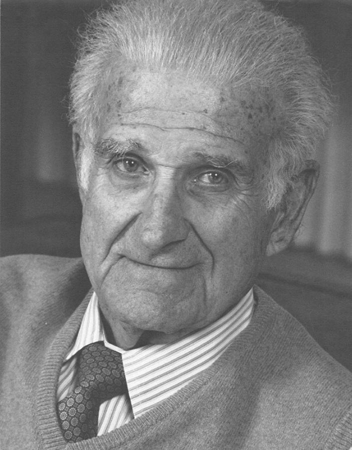 Black and white portrait of legendary flute maker George Koregelos. He is wearing an striped Oxford shirt and a patterned tie underneath a light-colored sweater vest. He is an elderly man with a gray, receding hairline. He is staring straight at the camera.