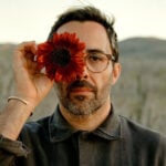 A bearded man in glasses holding a dark red sunflower over his right eye, posing against a blurred desert landscape in Mexico. He is Chef Paul Barbosa, who will be presenting a one-time pop-up dinner in Los Cabos, Baja California Sur.
