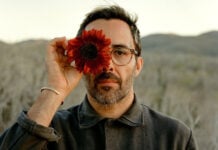 A bearded man in glasses holding a dark red sunflower over his right eye, posing against a blurred desert landscape in Mexico. He is Chef Paul Barbosa, who will be presenting a one-time pop-up dinner in Los Cabos, Baja California Sur.