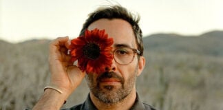 A bearded man in glasses holding a dark red sunflower over his right eye, posing against a blurred desert landscape in Mexico. He is Chef Paul Barbosa, who will be presenting a one-time pop-up dinner in Los Cabos, Baja California Sur.